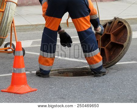 Worker In Uniform Stand Over The Open Sewer Hatch. Repair Of Sewage Or Underground Utilities, Nightm