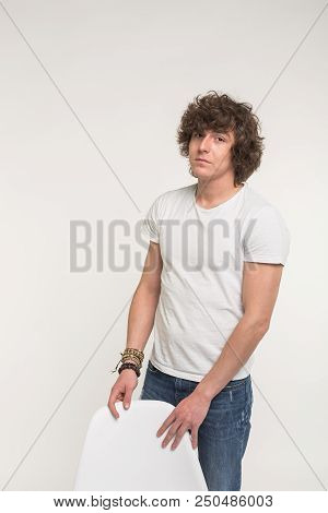 Handsome Young Man With Curly Hair Standing In White T-shirt Behind A Chair.