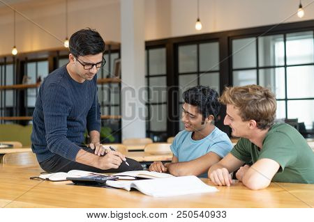 Serious Teacher Checking Assignment Of Two Students. Serious Man In Glasses Pointing Pen At Notes Of