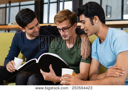 Three Fellow Students Reading Textbook And Drinking Coffee Together. Young Men Sitting On Couch. Edu