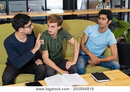 Three Fellow Students Studying And Discussing Homework. Young Men Sitting On Couch At Desk In Librar