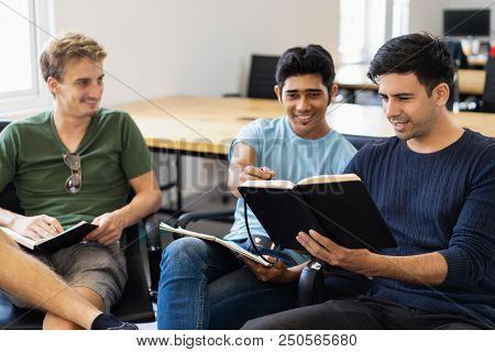 Three Fellow Students Studying, Reading Textbooks And Talking. Young Men Sitting In Armchairs In Cla