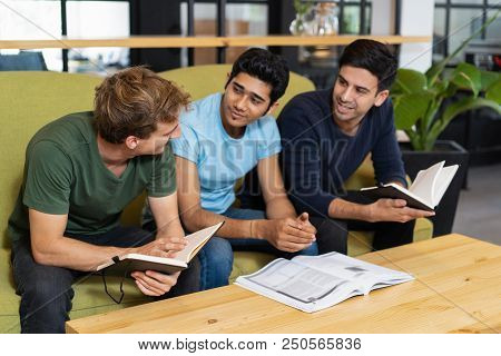 Three Fellow Students Talking And Studying Together. Young Men Sitting On Couch In Library Reading R