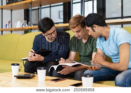 Three Focused Fellow Students Doing Homework Together. Young Men Sitting On Couch At Desk. Education