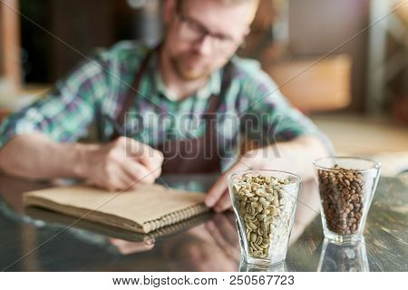 Two Glasses Of Coffee Beans, Raw And Roasted On Table In Modern Roastery With Blurred Form Of Barist