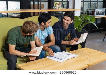 Three Relaxed Fellow Students Studying And Chatting. Young Men Sitting On Couch In Library Reading R