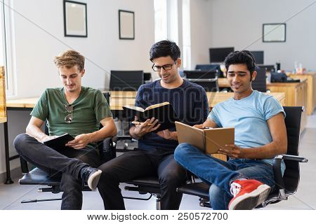 Three Smiling Fellow Students Studying And Reading Textbooks. Young Men Sitting In Armchairs In Clas
