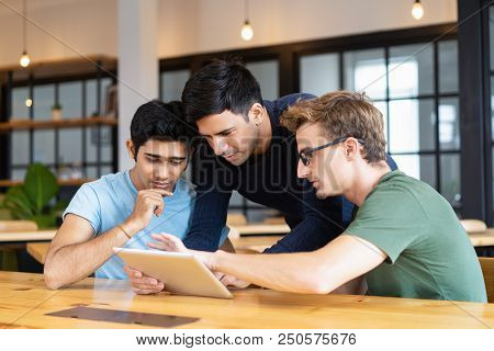 Three Thoughtful Students Using Tablet Computer. Young Men Sitting At Desk In Classroom Or Library. 