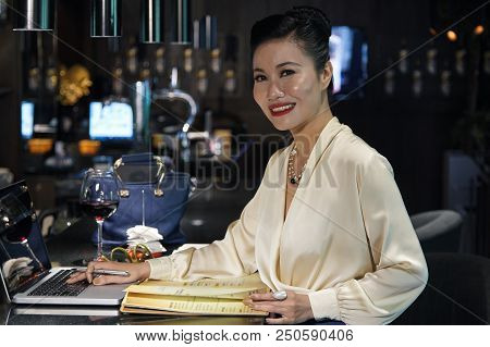 Side View Of Attractive Young Happy Asian Female In Light Elegant Blouse Using Laptop Placed On Dark