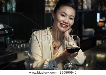 Relaxed Cheerful Asian Businesswoman In Elegant Blouse And Necklace Looking At Glass Of Red Wine Sta