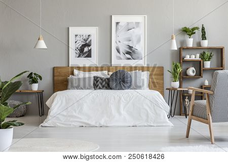 Posters And Lamps Above Wooden Bed In Grey Bedroom Interior With Patterned Armchair. Real Photo