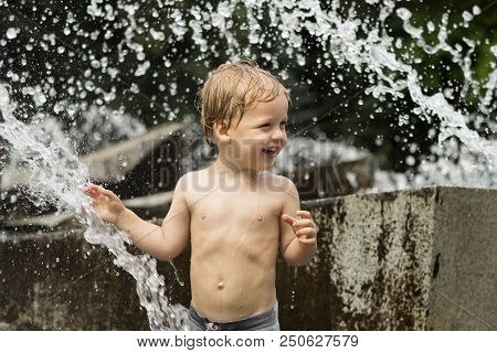 Funny Smiling Baby Boy Trying To Cauch Water Stream In Fountain. Cute Toddler Playing In The City Fo