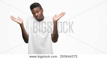 Young african american man wearing white t-shirt clueless and confused expression with arms and hands raised. Doubt concept.