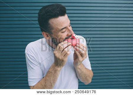Another Picture Of Man Biting A Piece Of Red Donut. He Likes Sweets. Isolated On Striped And Blue Ba