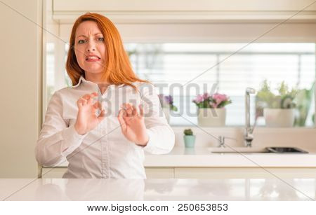 Redhead woman at kitchen disgusted expression, displeased and fearful doing disgust face because aversion reaction. With hands raised. Annoying concept.
