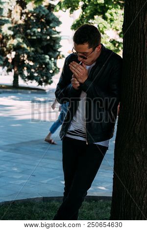 A Young Guy Smokes Near A Tree In The City. A Black Jacket, Jeans, White Sneakers, A White T-shirt A