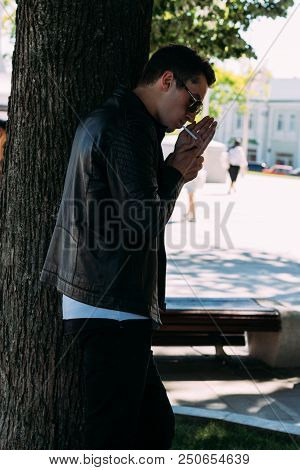 A Young Guy Smokes Near A Tree In The City. A Black Jacket, Jeans, White Sneakers, A White T-shirt A