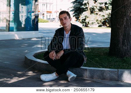 A Young Guy Smokes Near A Tree In The City. A Black Jacket, Jeans, White Sneakers, A White T-shirt A