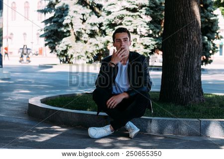 A Young Guy Smokes Near A Tree In The City. A Black Jacket, Jeans, White Sneakers, A White T-shirt A