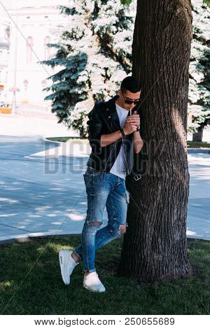 A Young Guy Smokes Near A Tree In The City. A Black Jacket, Jeans, White Sneakers, A White T-shirt A