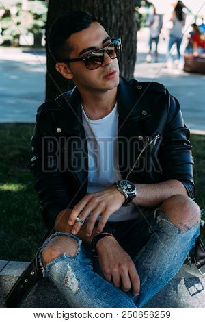 A Young Guy Smokes Near A Tree In The City. A Black Jacket, Jeans, White Sneakers, A White T-shirt A