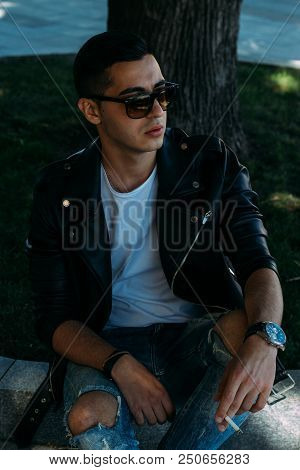 A Young Guy Smokes Near A Tree In The City. A Black Jacket, Jeans, White Sneakers, A White T-shirt A