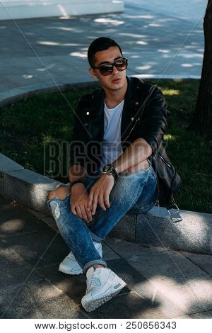 A Young Guy Smokes Near A Tree In The City. A Black Jacket, Jeans, White Sneakers, A White T-shirt A