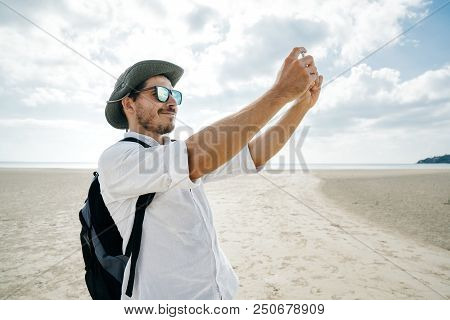 Tourist In A Hat With Glasses With A Backpack Is Holding A Smartphone In His Hands And Is Making A P