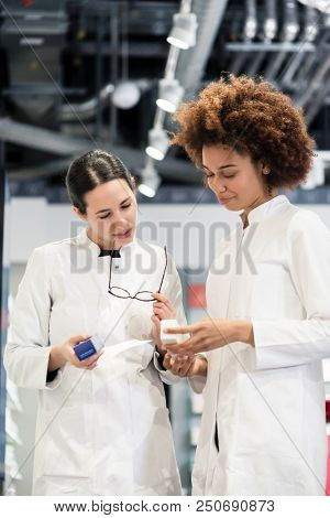Low-angle view of two reliable pharmacists analyzing together a medical prescription and two different packages of medicine during work in a modern drugstore