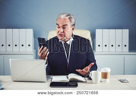 Shocked Angry Senior Male In Elegant Business Suit Sitting At Work Desk With Notepad And Gadgets And