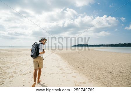 A Free-traveled Man On A Tropical Beach Barefoot With A Backpack In A White Shirt, Hat, Shorts And G
