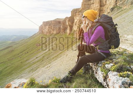 Smiling Girl Traveler In A Yellow Hat And A Pair Of Sunglasses Ssitting At The Foot Of Epic Rocks Wi