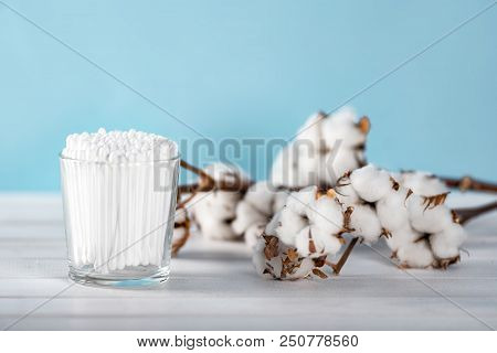 Cotton Swabs In Clear Glass Jar In Bathroom Setting With A Cotton Flowers On Pastel Blue Background.