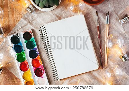 Top View Mockup With Notepad, Paints, Brushes, Flower, Dummy And Bulb On Wood And Fabric Background.