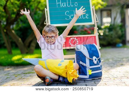 Happy Little Kid Boy With Glasses Sitting By Desk And Backpack Or Satchel. Schoolkid With Traditiona