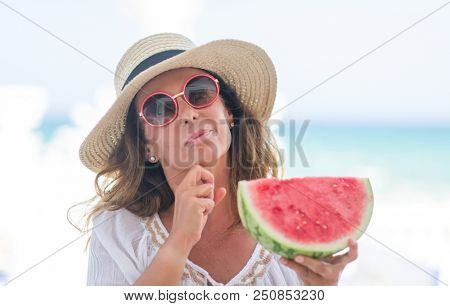 Middle age brunette woman by the sea eating watermelon serious face thinking about question, very confused idea