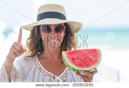Middle age brunette woman by the sea eating watermelon surprised with an idea or question pointing finger with happy face, number one