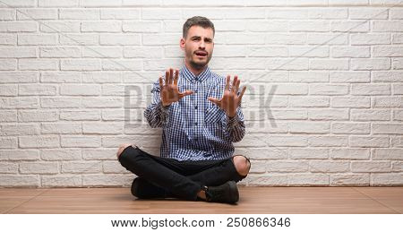 Young adult man sitting over white brick wall afraid and terrified with fear expression stop gesture with hands, shouting in shock. Panic concept.
