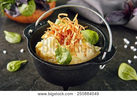 Mashed Potatoe Decorated with Roasted Onions and Greens in Cast Iron Pot Close Up. Creamy Whipped Potatoes Puree with Butter on Selective Focus Blurred Background