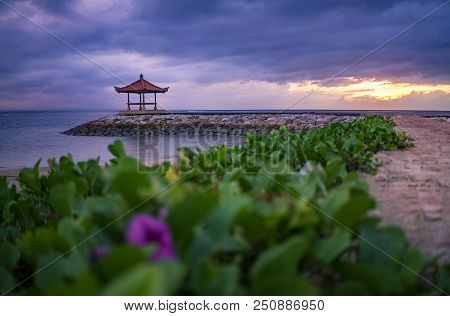 Sunrise In Cloudy Morning At Sanur Beach With Pavilion And Ornamental Plant. A View From Hotel Garde