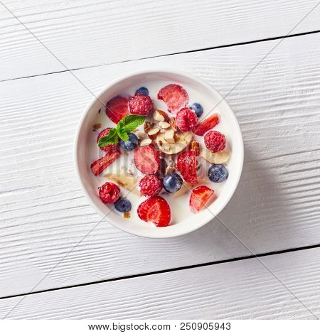 Colorful and tasty ready breakfast from corn muesli, berries, milk, almonds, slices banana on a bowl on a white wooden table with copy space. Flat lay.