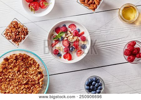 Homemade granola in a plate, sliced banana, berries, almonds, walnuts and white bowl with natural organic breakfast on white wooden table.