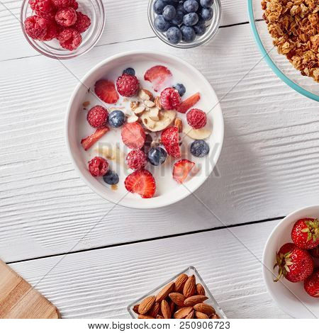 Ingredirnts for preparing healthy organic breakfast - granola, strawberries, honey, banana in white bowl with milk on white table. Flat lay.