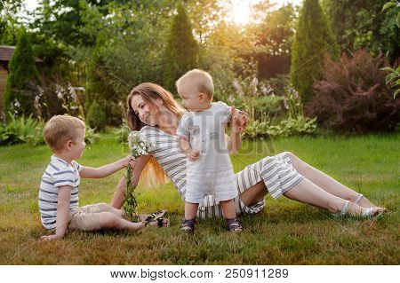 Mom, Two Children Rest On Nature. Sibling Rivalry. Brothers, The Son Gives His Mother Flowers. Mothe