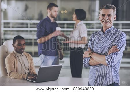 Smiling Booss And His Team Is Working In Office. Chief With Crossed Hands. Business People On Backgr
