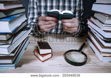 A Man Reading A Miniature Book Among The Big Books In The Library At The Wooden Table.