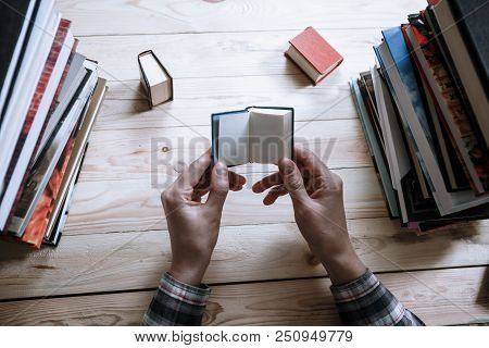 A Man Reading A Miniature Book Among The Big Books In The Library At The Wooden Table.
