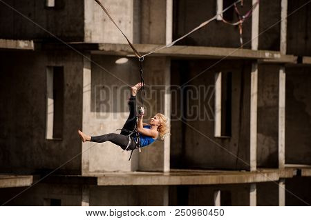 Young Female Equilibrist Hanging On The Slackline Upside Down On The Background Of Unfinished Buildi