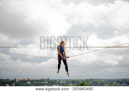 Female Equilibrist Sitting On The Slackline Rope On The Background Of Houses Among Trees And Clear B