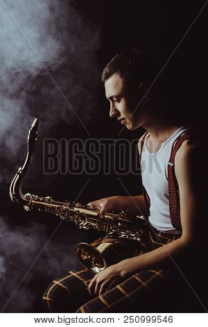 Side View Of Stylish Young Jazzman Sitting And Holding Saxophone In Smoke On Black